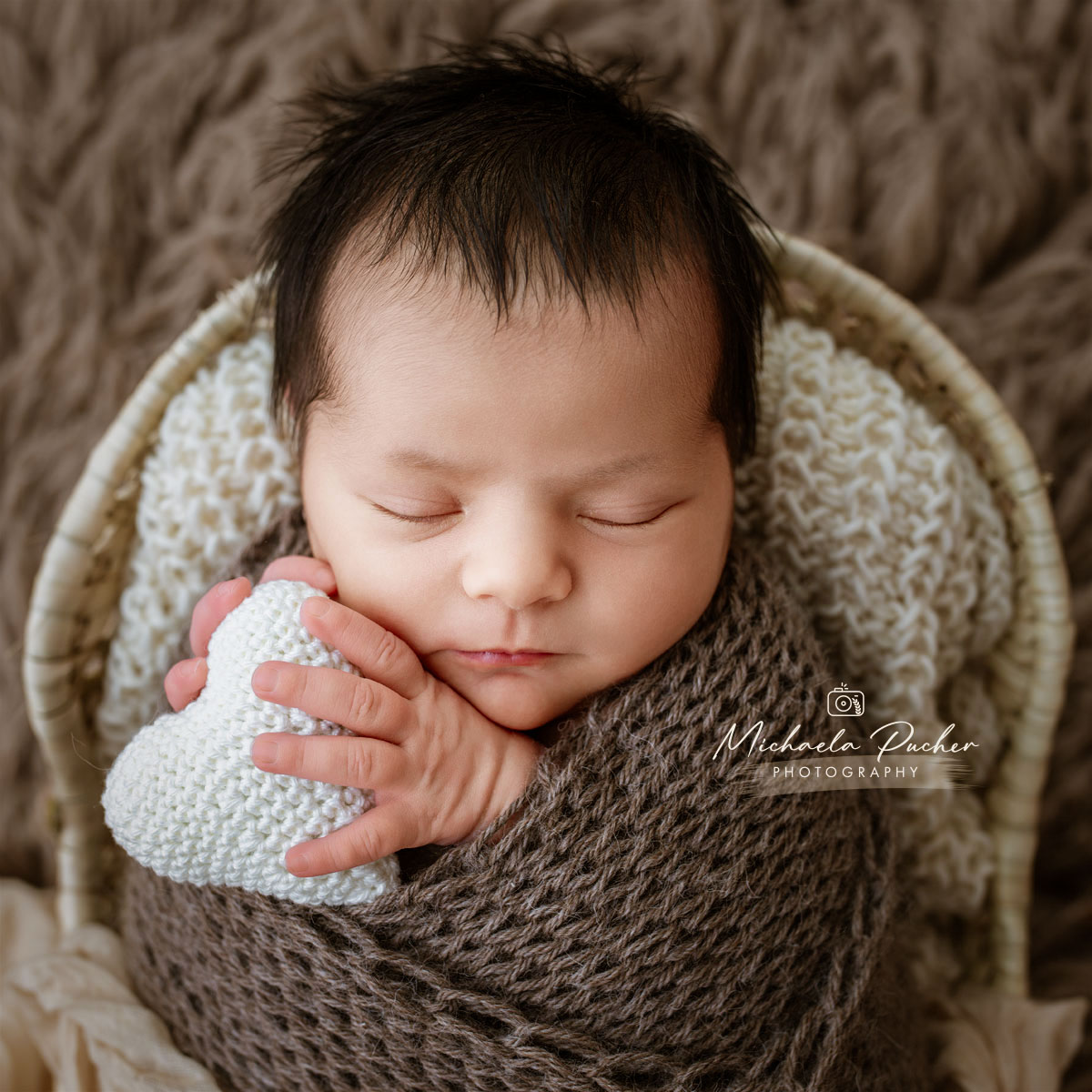 Schlafendes Neugeborenes, eingewickelt in braune Wolle, hält ein gehäkeltes weißes Herz in den Händen und liegt auf weichen Decken.